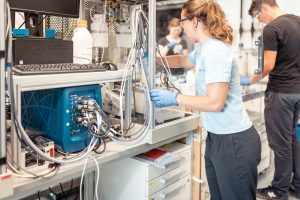 A laboratory image containing parts of an electrolysis test bench and a scientist reaching across the cell fixture.
